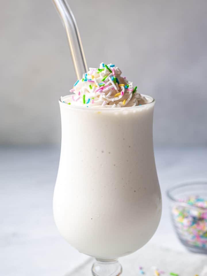 A vegan vanilla milkshake in a hurricane glass topped with non-dairy whipped cream and sprinkles.
