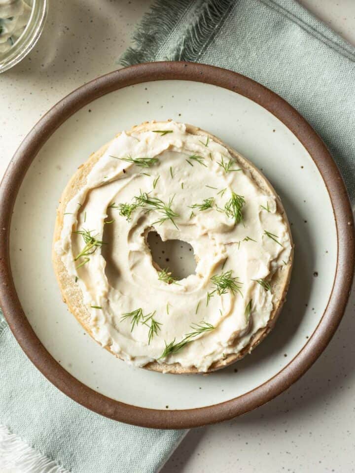 Half of a bagel spread with tofu cream cheese and sprinkled with dill resting on a small plate.