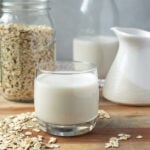 A glass of oat milk with a jar of oats and a tall jar of oat milk in the background.