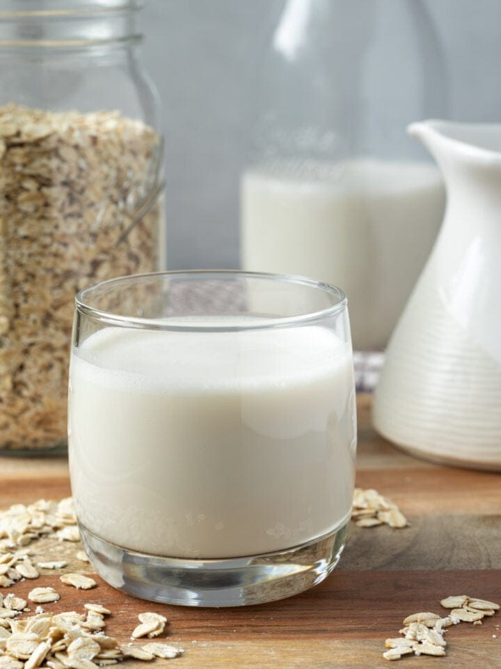 A glass of oat milk with a jar of oats and a tall jar of oat milk in the background.