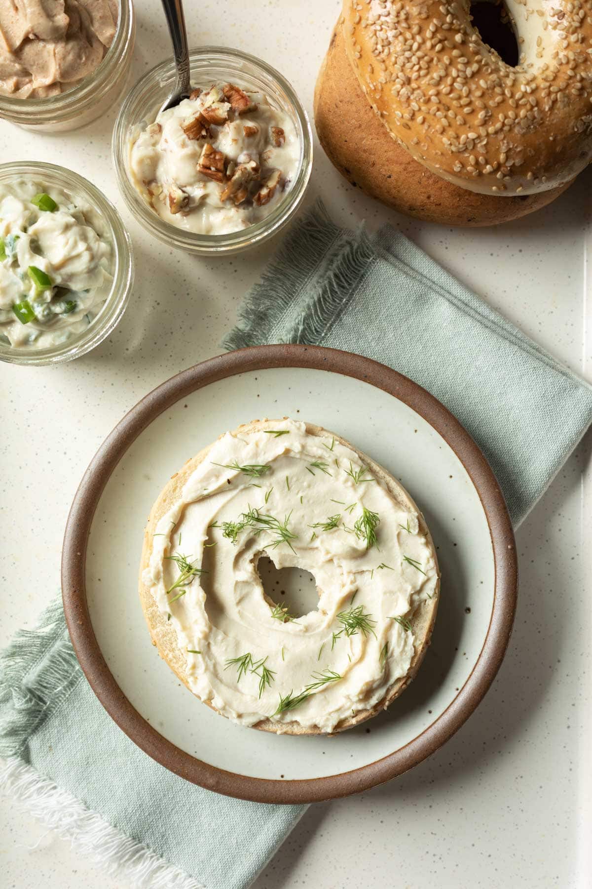 Tofu-oat cream cheese on half a bagel with more bagels and flavored vegan cream cheese in background.