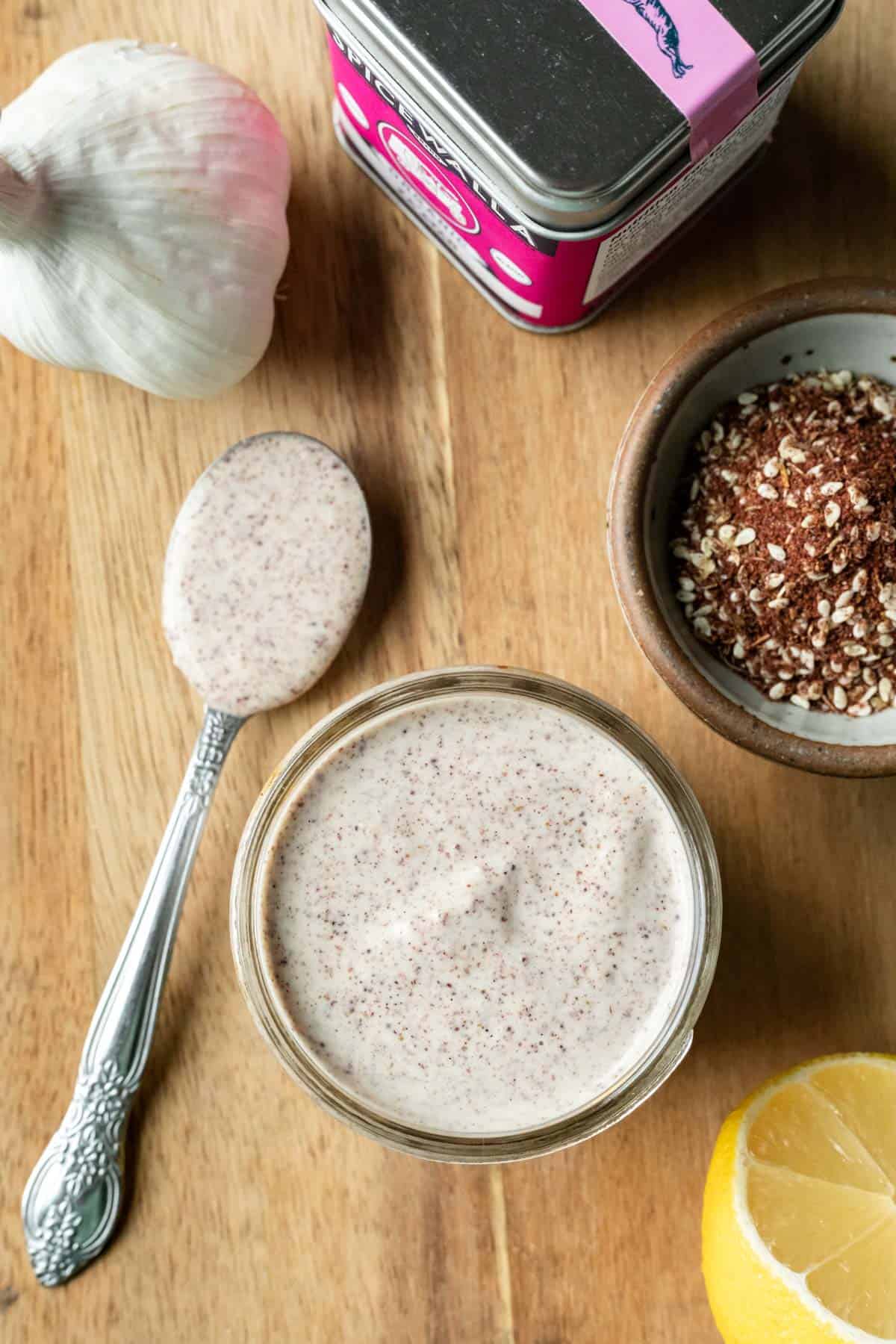 A jar of creamy za'atar dressing on a wooden board with lemon and dry seasoning nearby.