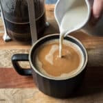 A hand pouring oat creamer into a black mug filled with coffee.