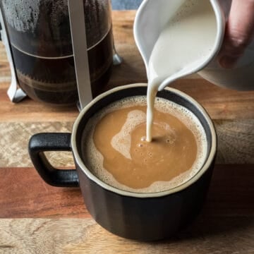 A hand pouring oat creamer into a black mug filled with coffee.