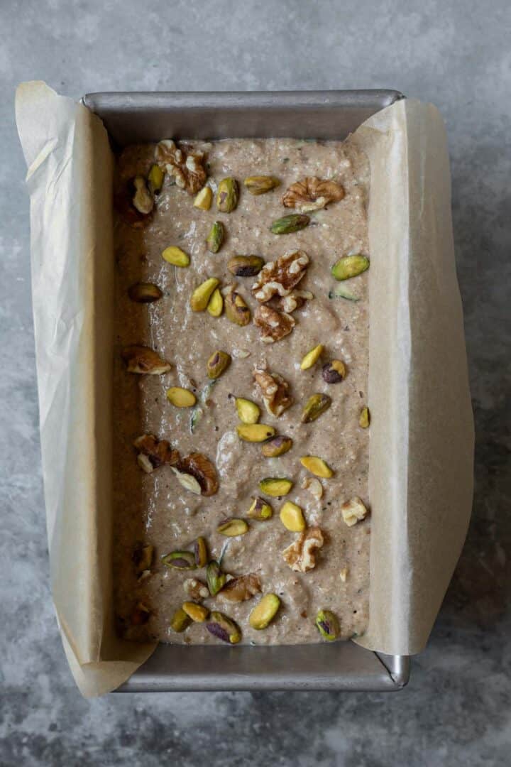 Batter in a parchment-lined loaf pan that has walnuts and pistachios sprinkled on top.