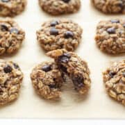 banana cookies with chocolate chips on a baking sheet with one cookie broken in half to show texture.