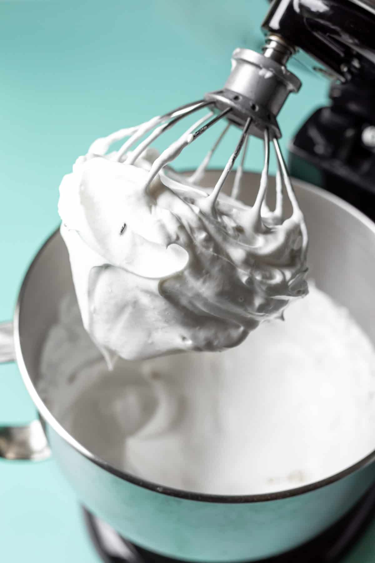 Vegan whipped cream clinging to the whisk of a mixer against an aqua blue background.