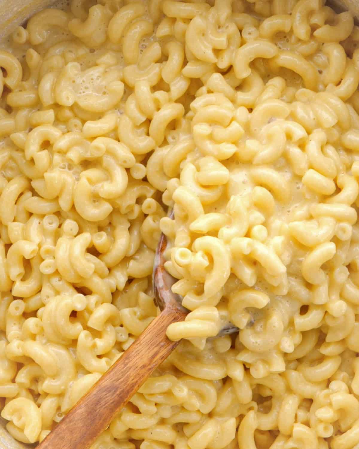 Close up of macaroni with extra creamy cheese sauce being scooped up with a large wooden spoon.