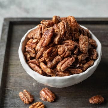 Crisp candied pecans in a small white bowl resting on a dark wood serving board.
