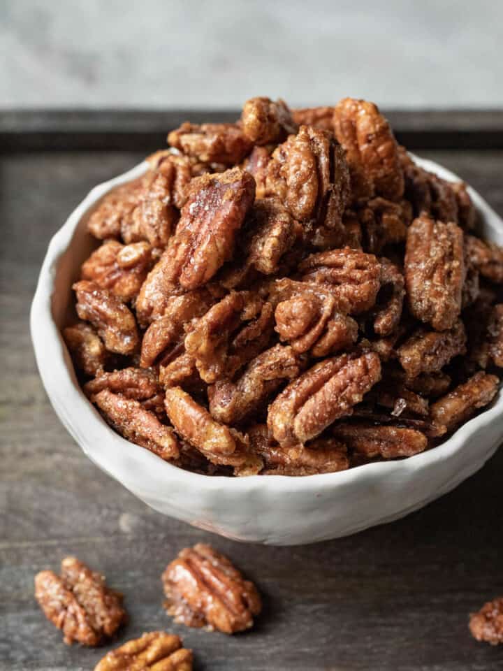 Crisp candied pecans in a small white bowl resting on a dark wood serving board.