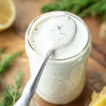 A glass jar filled with creamy salad dressing with lemons and fresh dill on a cutting board.
