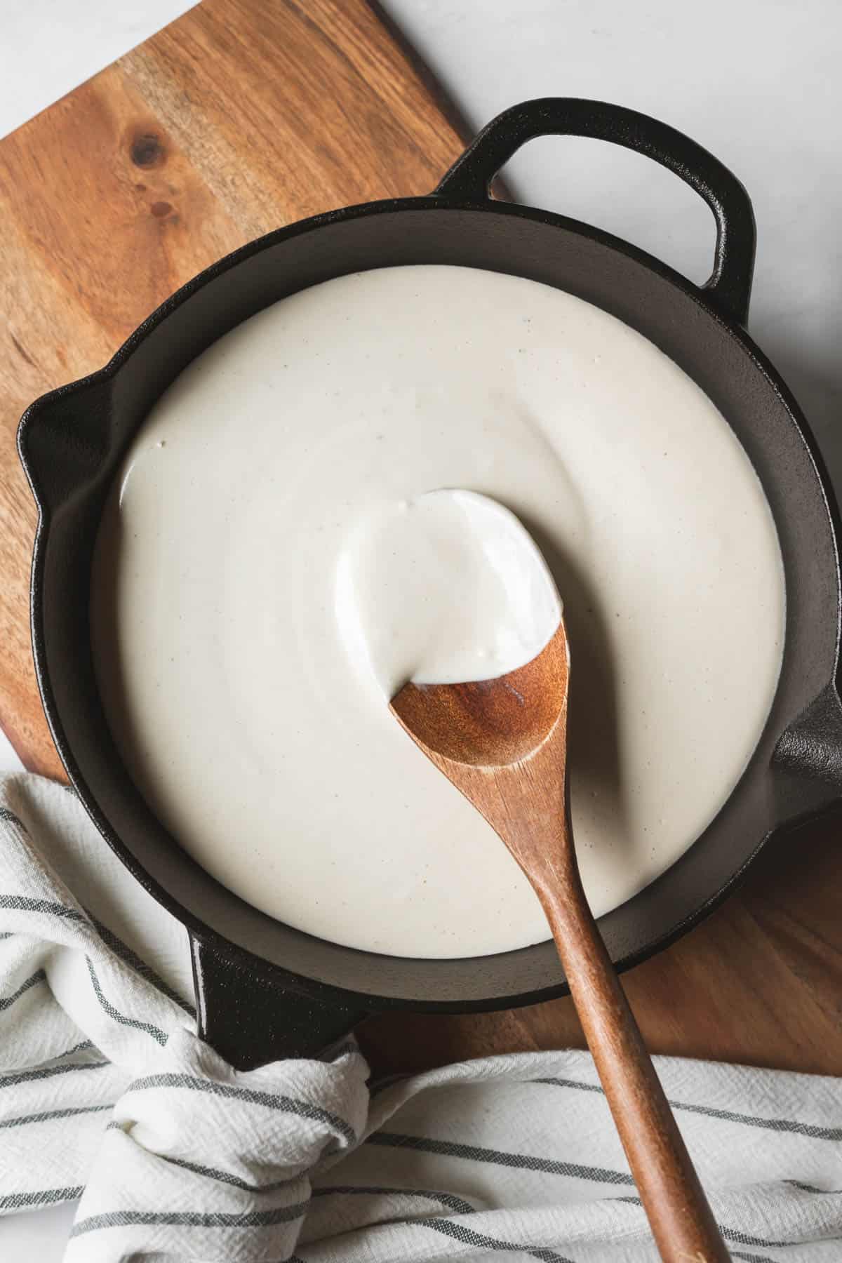 A cast iron pan filled with creamy vegan bechamel stirred by a wooden spoon.