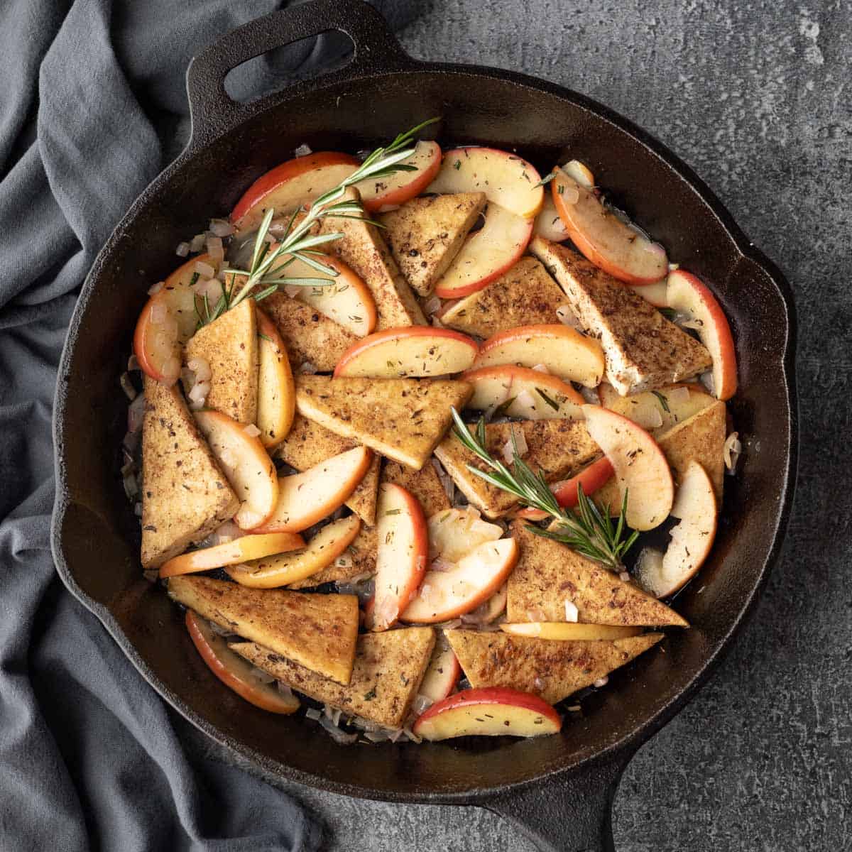 A large skillet filled with tofu, apples, rosemary, and shallot, and glazed with apple cider.