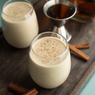 Two glasses of vegan oat nog on a wood board with a shot of bourbon in background.