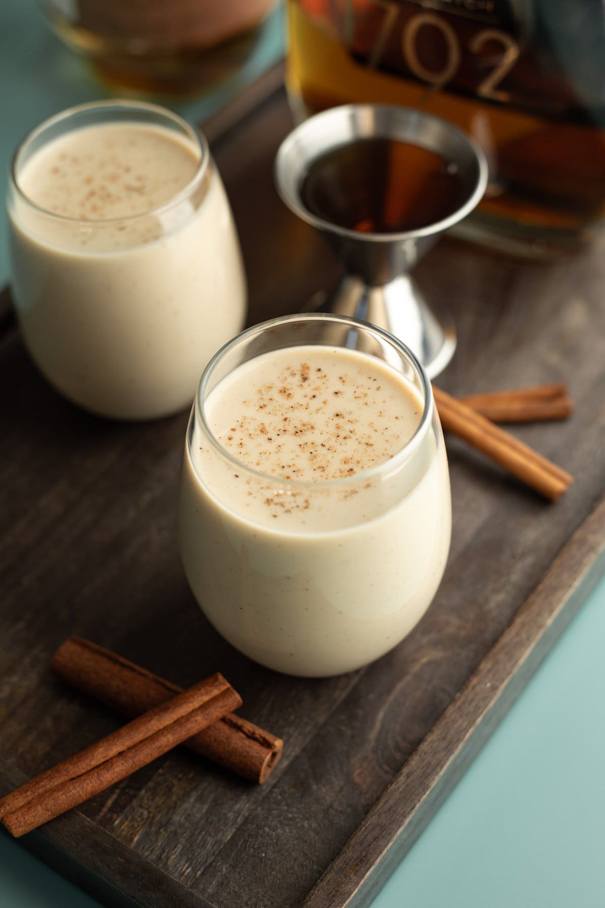 A dark wood board with 2 glasses of vegan eggnog and a shot of bourbon.