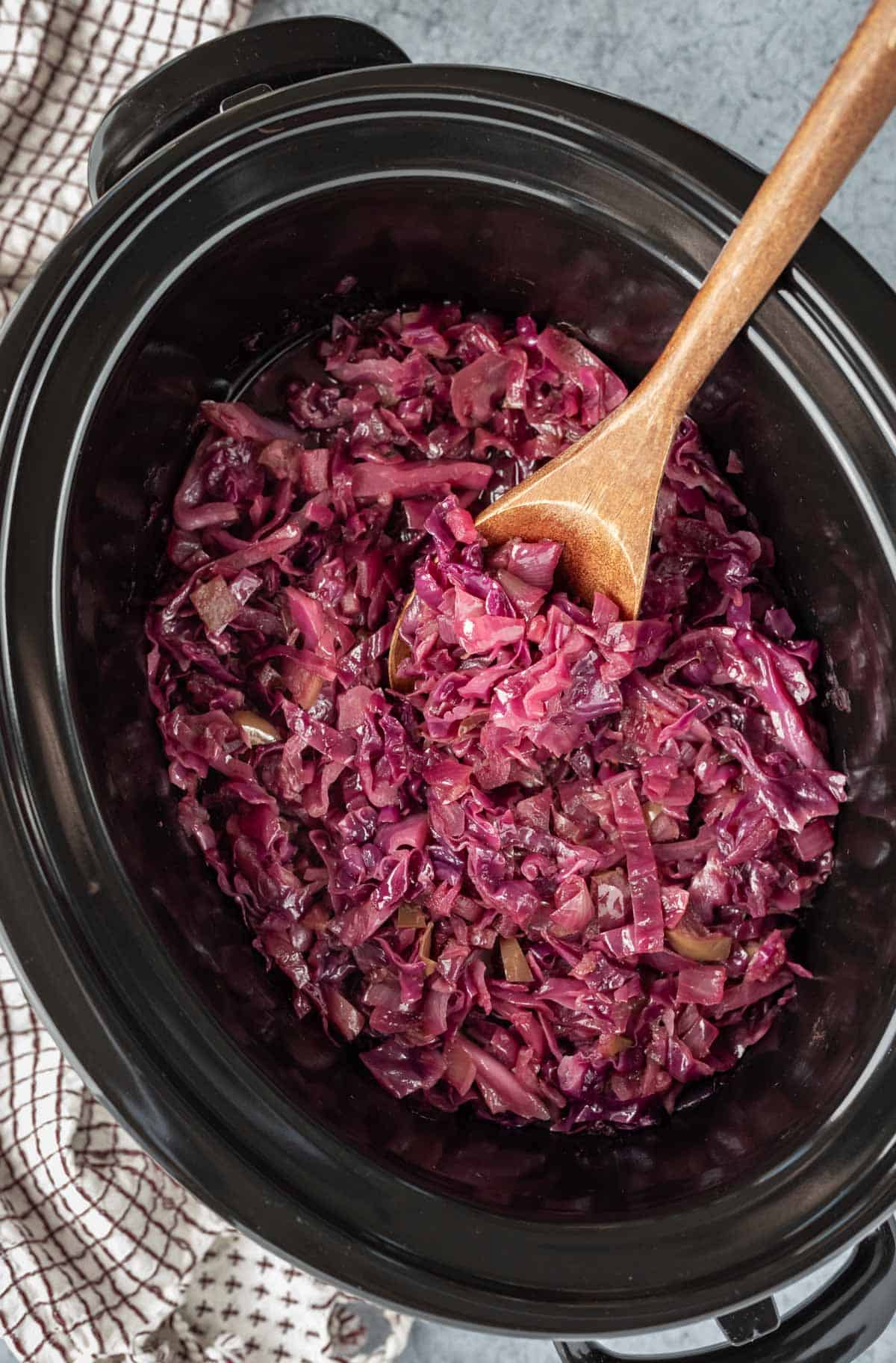 Cooked German red cabbage in a slow cooker with a wooden serving spoon resting on the side.