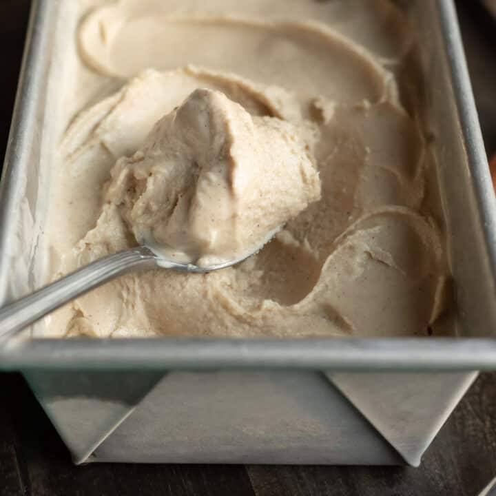 A spoon scooping up creamy, melting eggnog ice cream from a metal loaf pan.