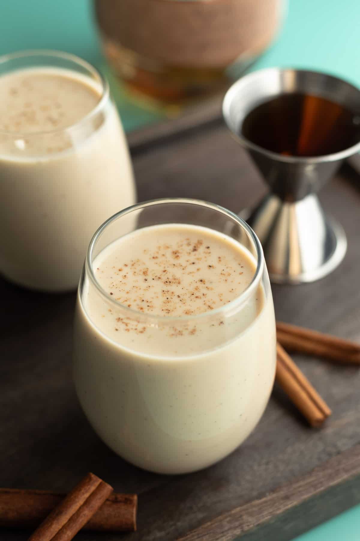 A short glass filled with oat milk eggnog with cinnamon sticks and bourbon nearby.