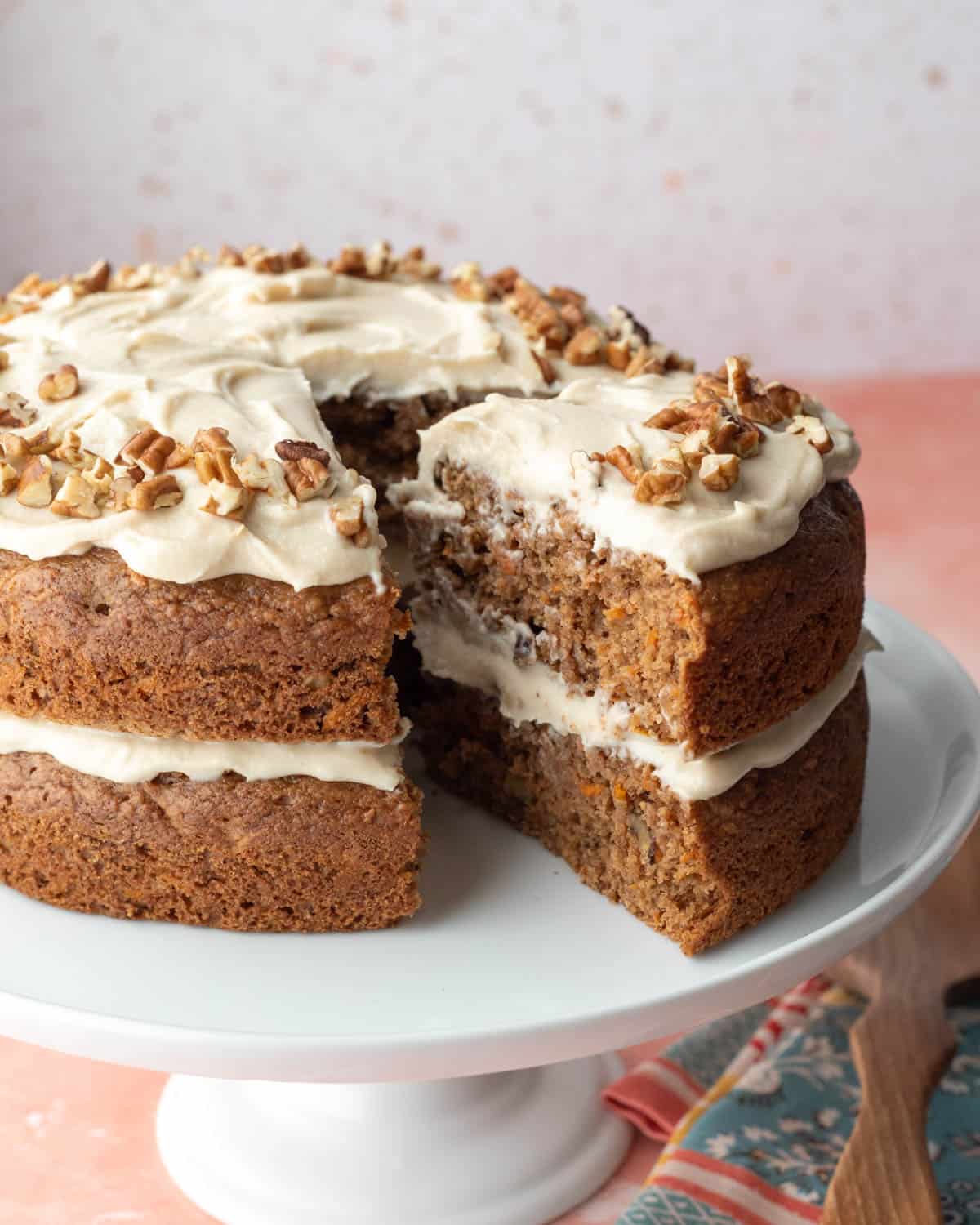 Two-layer vegan carrot cake on a white cake stand with a slice pulled out to show texture.