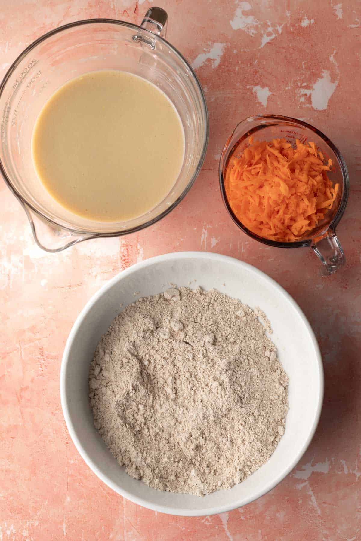 Two large mixing bowls filled with wet and dry ingredients plus a measuring cup with grated carrot.