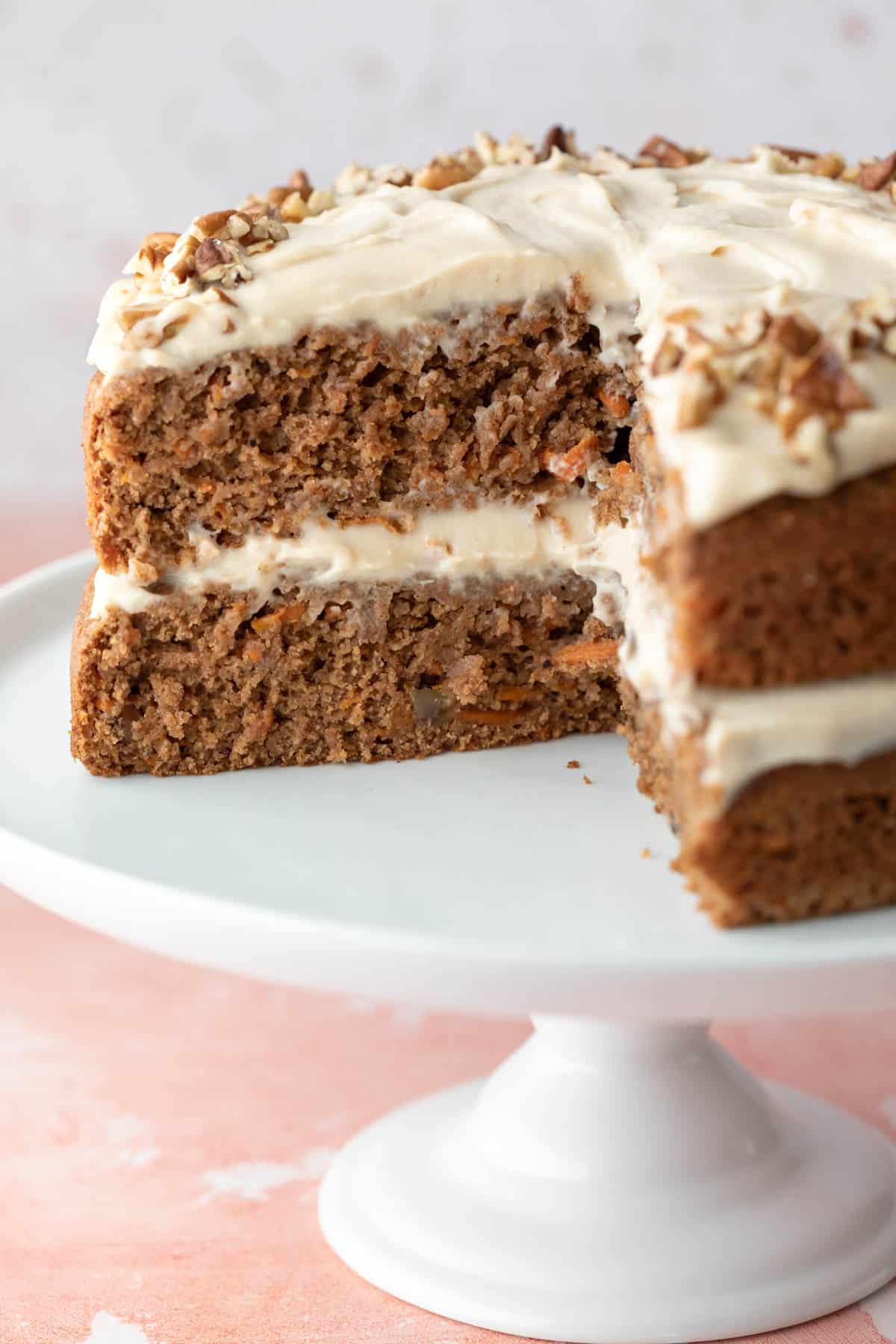 Carrot cake on a white stand with a quarter of the cake removed showing the frosting between the moist and fluffy layers.