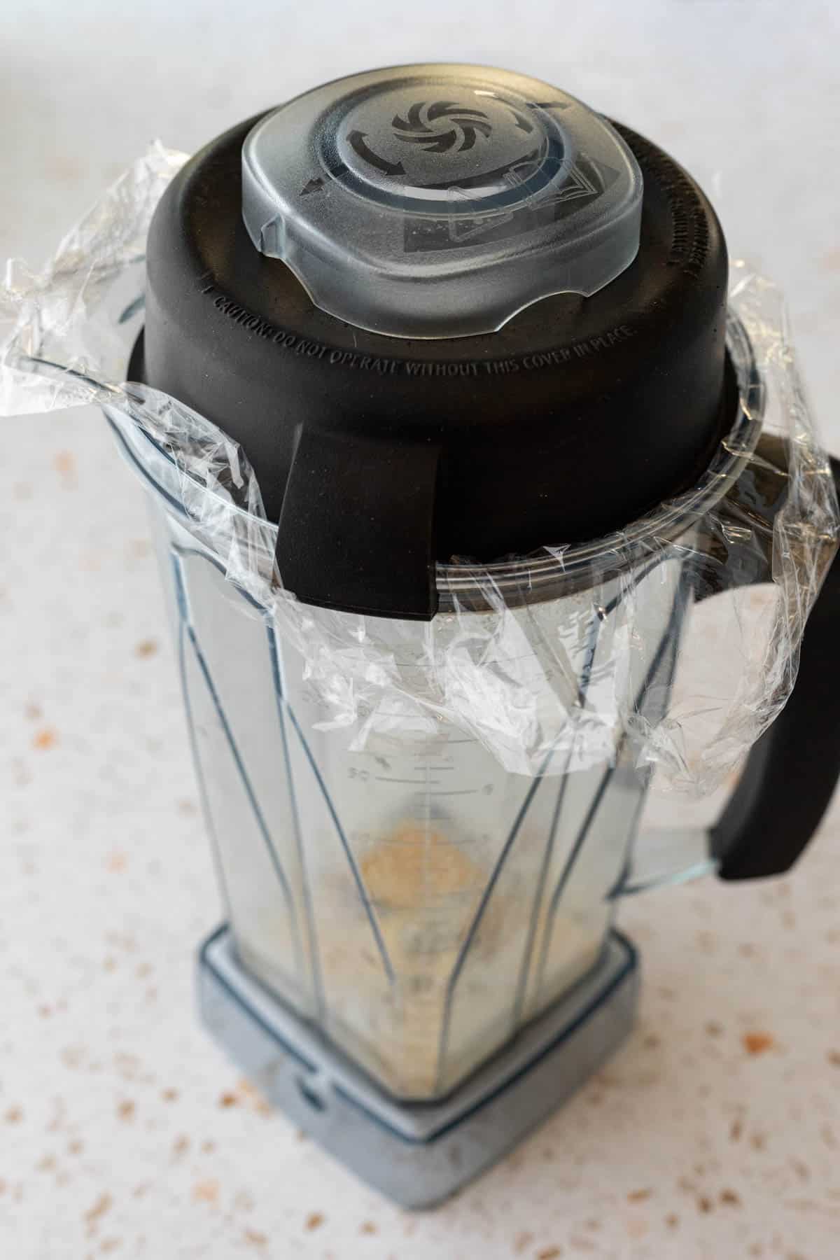 Dry polenta in a blender with plastic wrap underneath the lid.