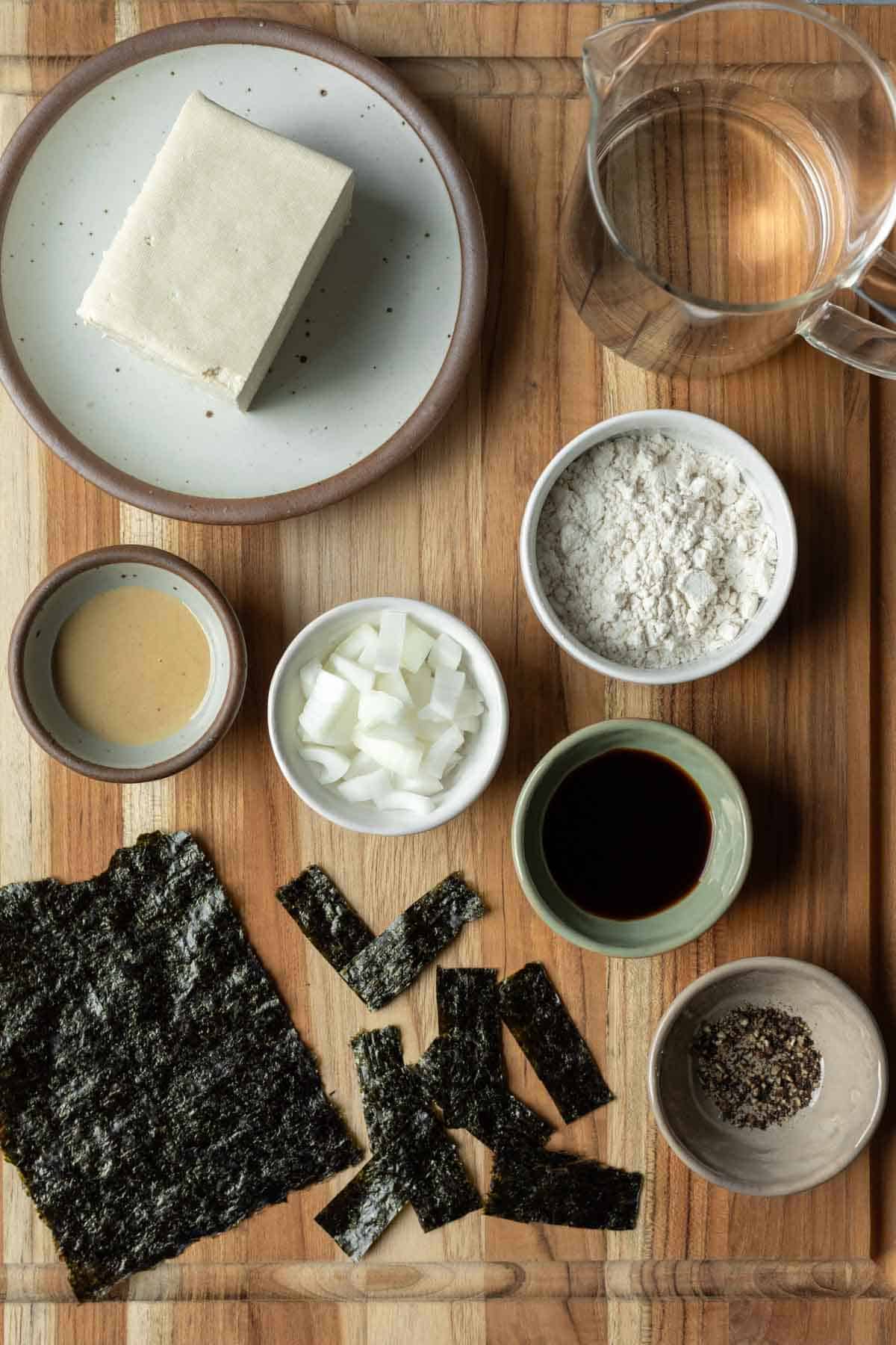 The 7 ingredients needed to make savory tofu pancakes laid out on a wood board.