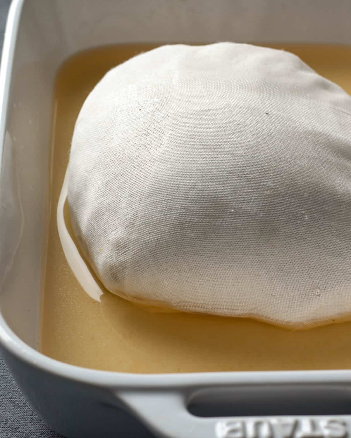 Wrapped vegan turkey resting in broth in a baking pan.