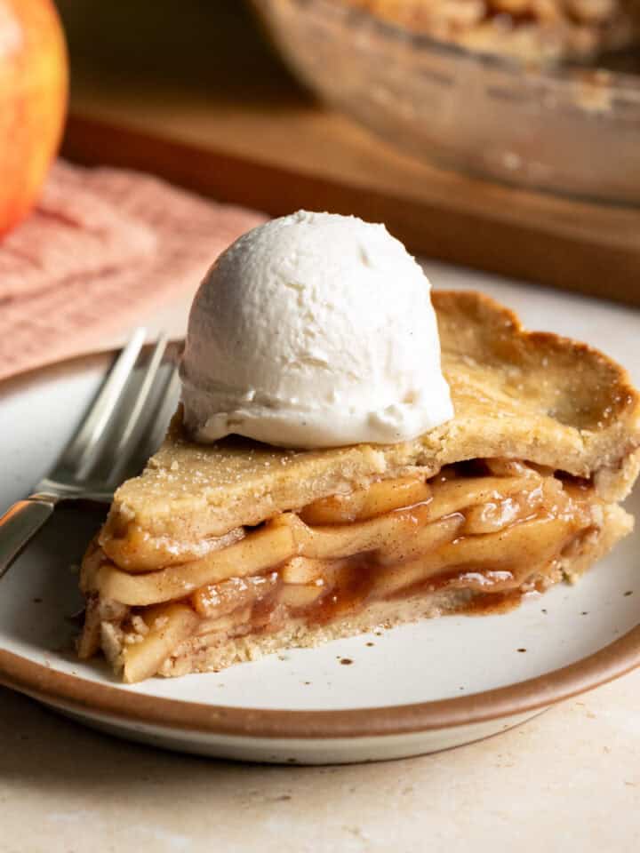 A slice of vegan apple pie topped with a scoop of vanilla ice cream on a small plate.