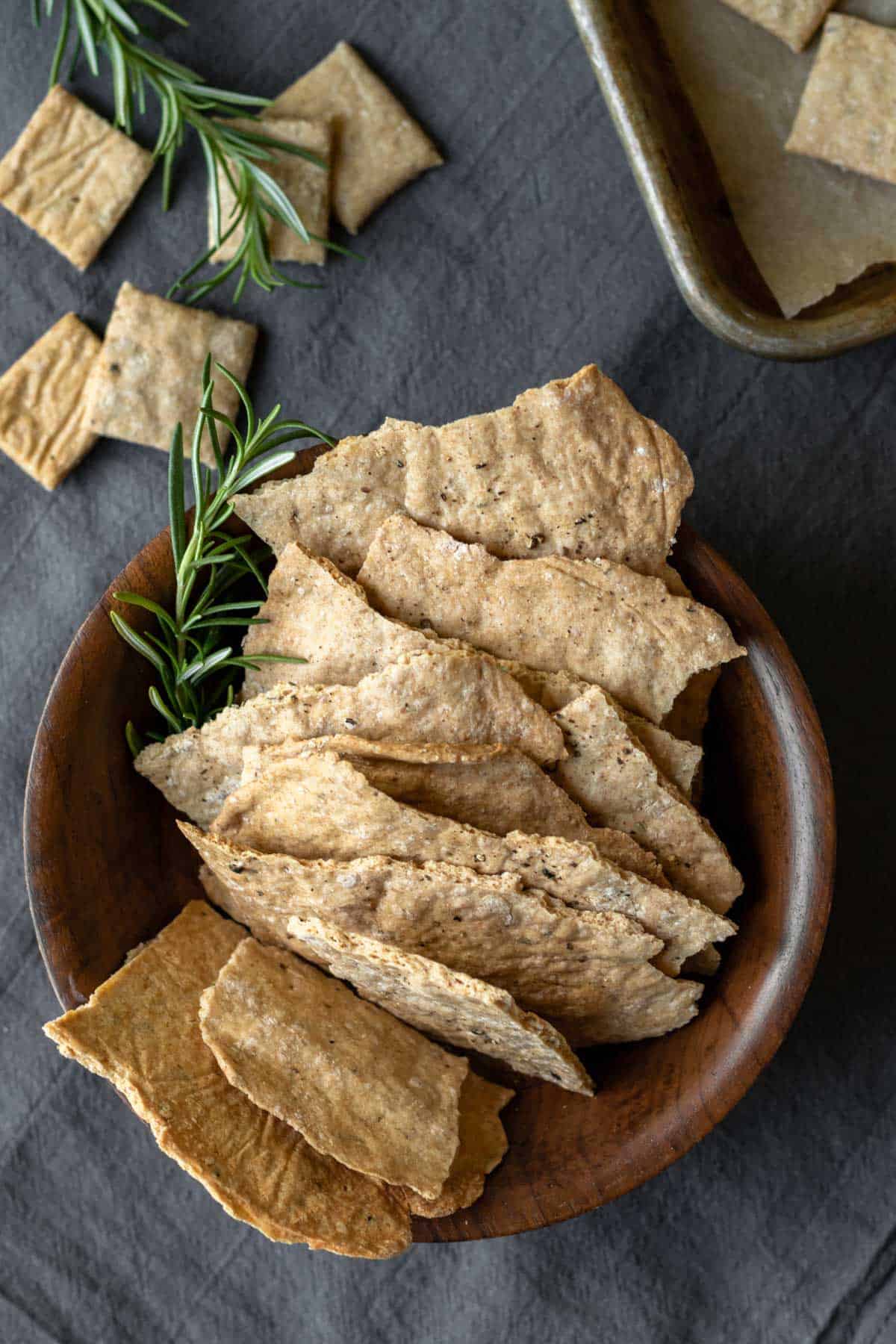 Artisan whole wheat crackers in a wooden bowl with rosemary garnish.