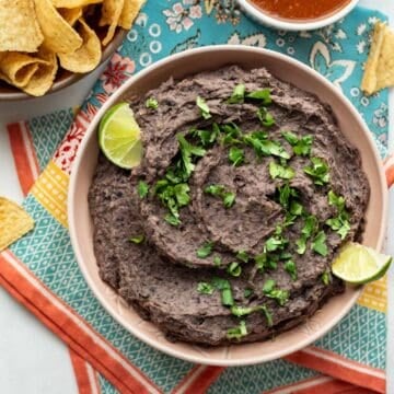 Close-up of refried black beans in a bowl with lime wedges and cilantro as garnish.