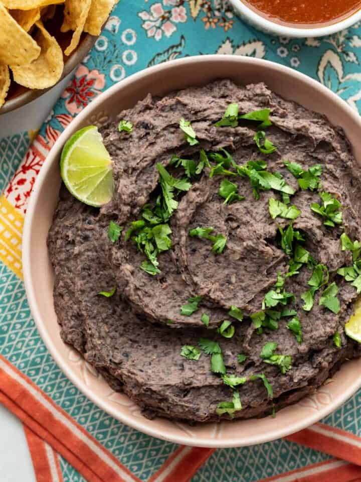 Close-up of refried black beans in a bowl with lime wedges and cilantro as garnish.