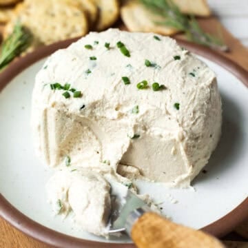 A creamy wheel of cashew Boursin cheese on a small plate with a cheese knife on the side.