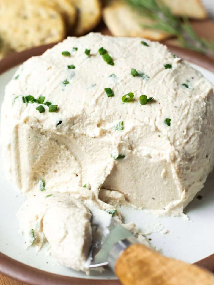 A creamy wheel of cashew Boursin cheese on a small plate with a cheese knife on the side.