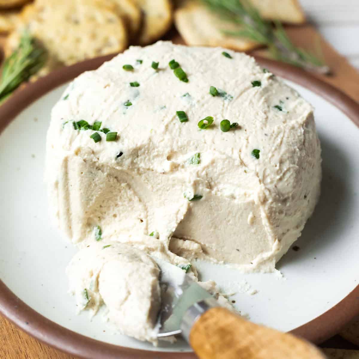 A creamy wheel of cashew Boursin cheese on a small plate with a cheese knife on the side.