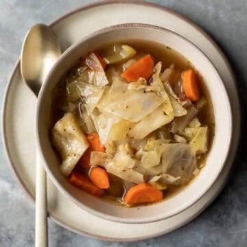 A serving of crockpot cabbage soup in a bowl set on a plate with a spoon next to it.