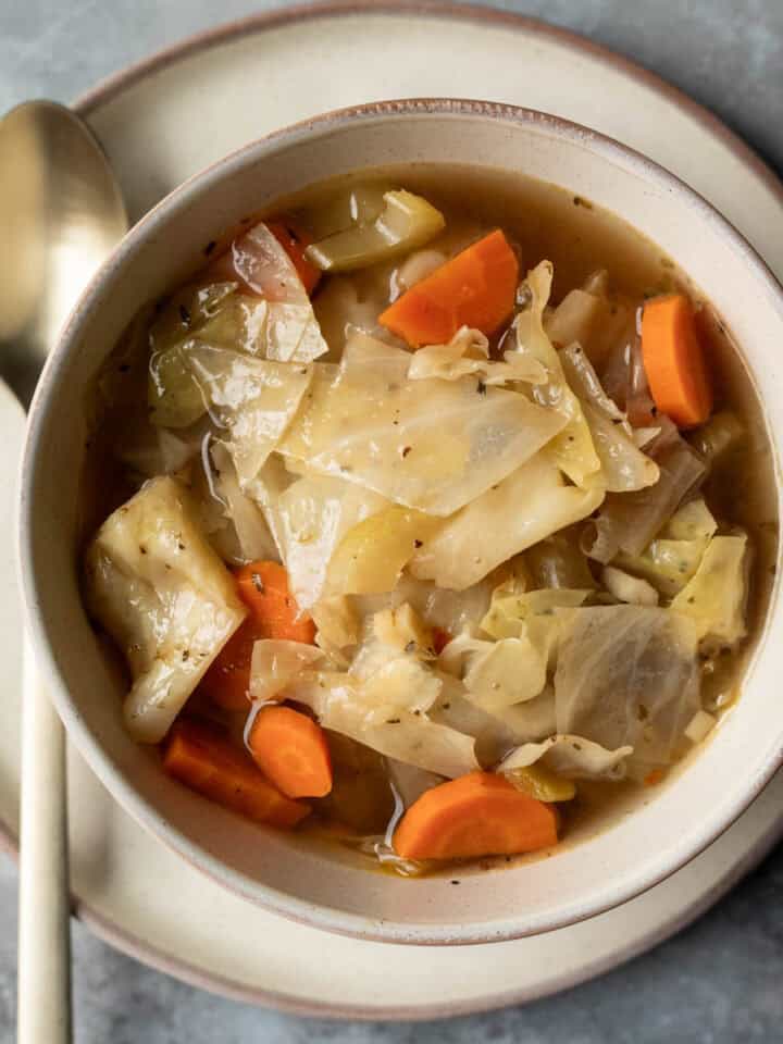 A serving of crockpot cabbage soup in a bowl set on a plate with a spoon next to it.