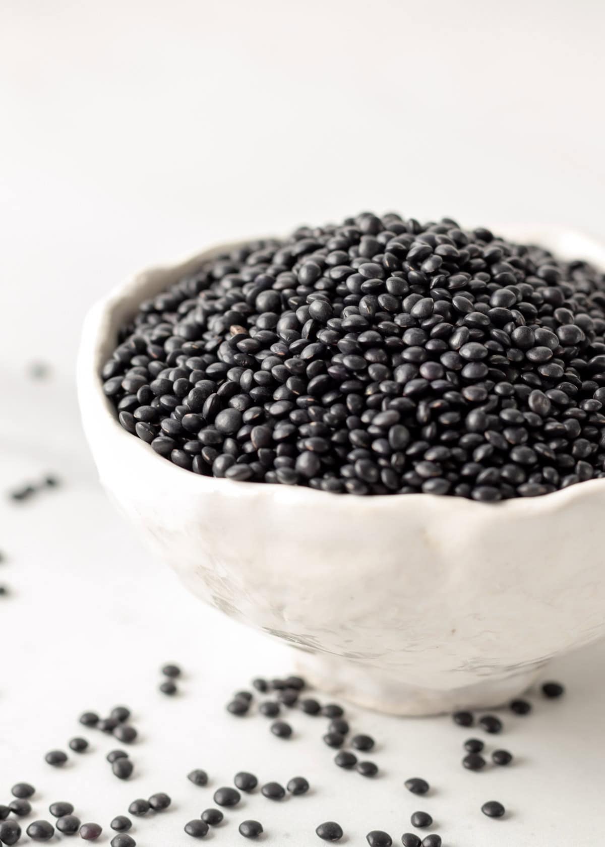 A white bowl filled with dry black lentils and some scattered on the table.