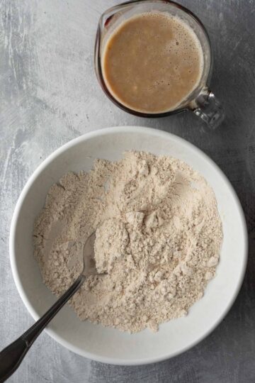 Dry ingredients for muffin batter in a white bowl with a spoon with the wet ingredient mixture in a measuring cup behind it.