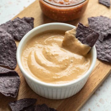 A white bowl of tofu queso with a blue tortilla chip dipped in it.