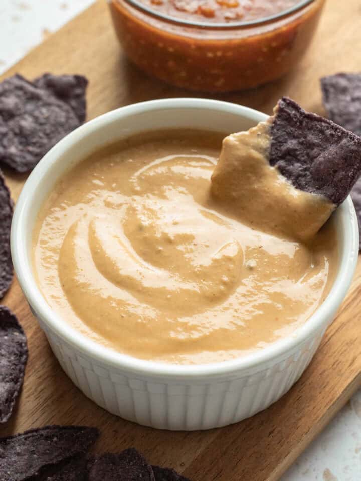 A white bowl of tofu queso with a blue tortilla chip dipped in it.