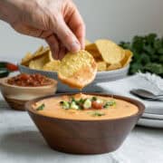 A hand holding a tortilla chip scooping up creamy vegan queso from a wood bowl.