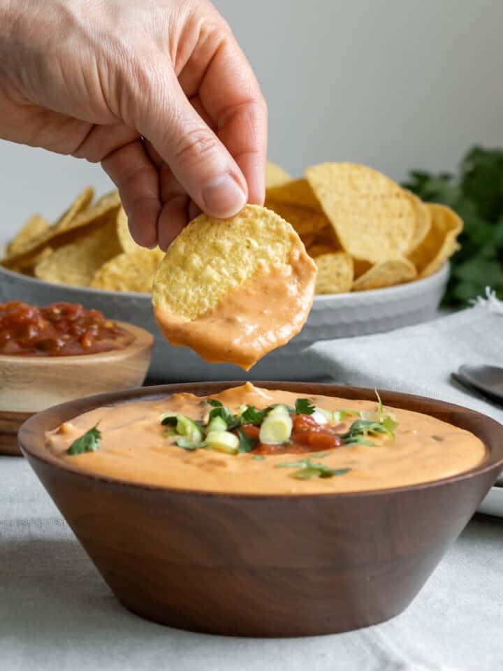 A hand holding a tortilla chip scooping up creamy vegan queso from a wood bowl.