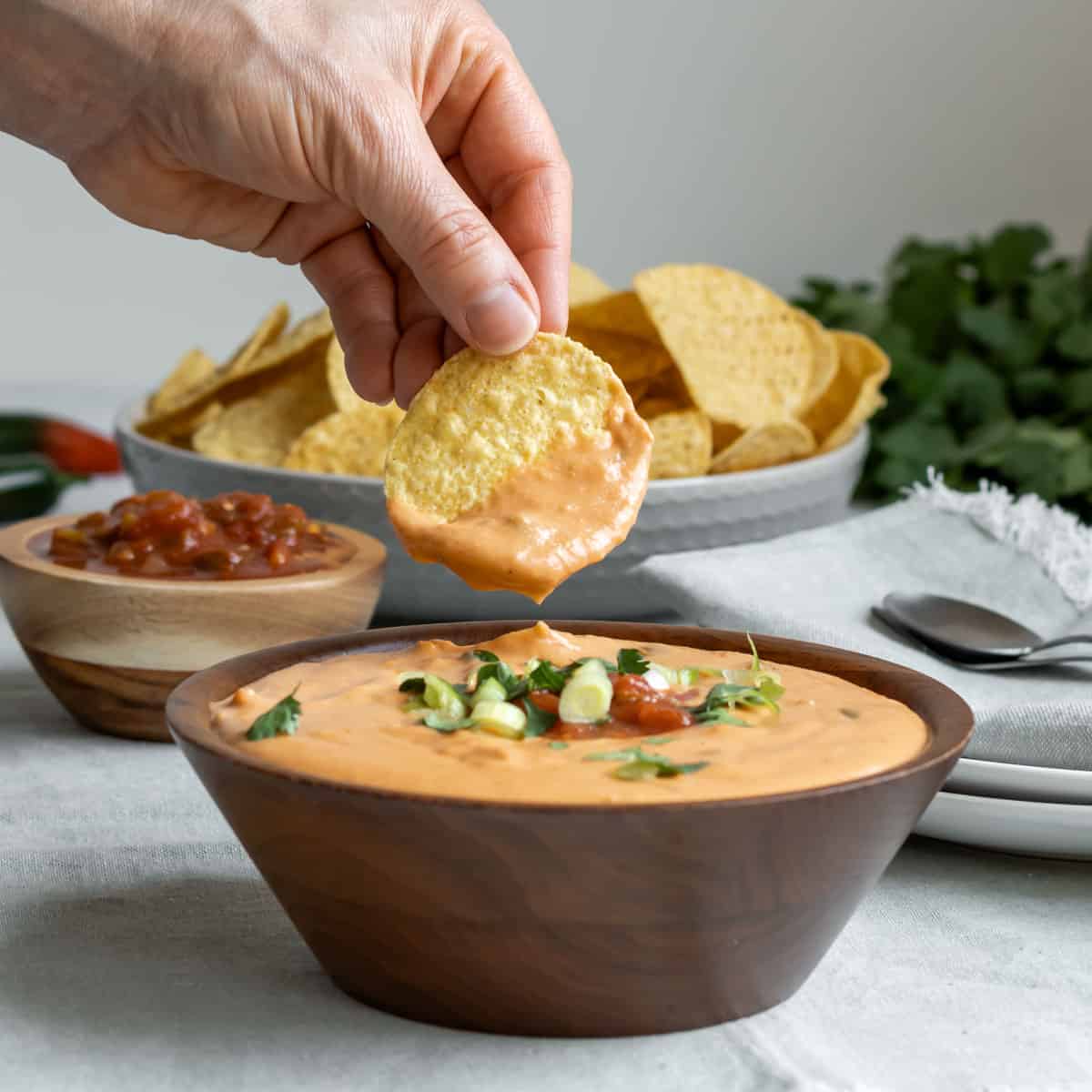 A hand holding a tortilla chip scooping up creamy vegan queso from a wood bowl.
