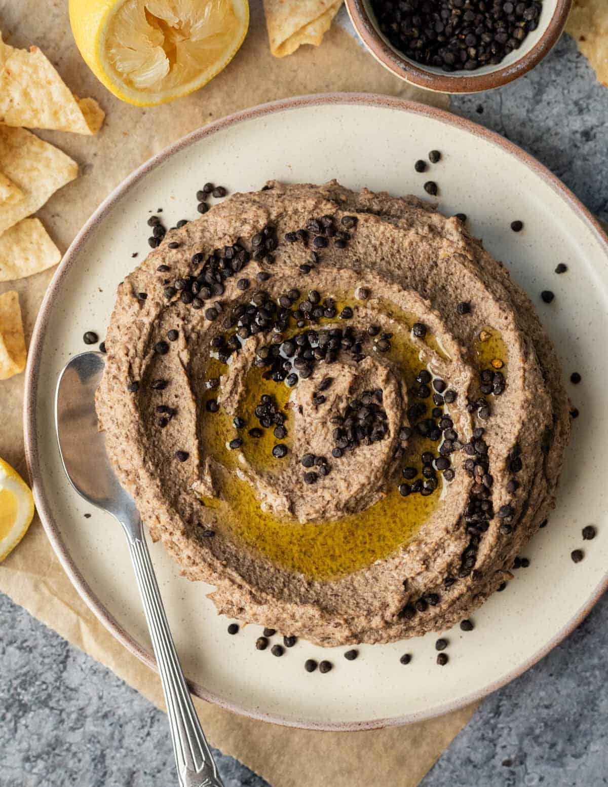 Black lentil hummus dip on a white serving plate topped with crispy lentils and olive oil.