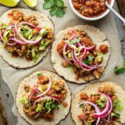 Four tempeh tacos laid out on a tray topped with pickled red onion.