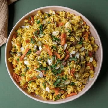 Colorful brown rice pilaf with nuts, seeds, and apricots in a bowl on a green surface.