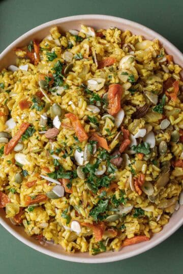 Colorful brown rice pilaf with nuts, seeds, and apricots in a bowl on a green surface.