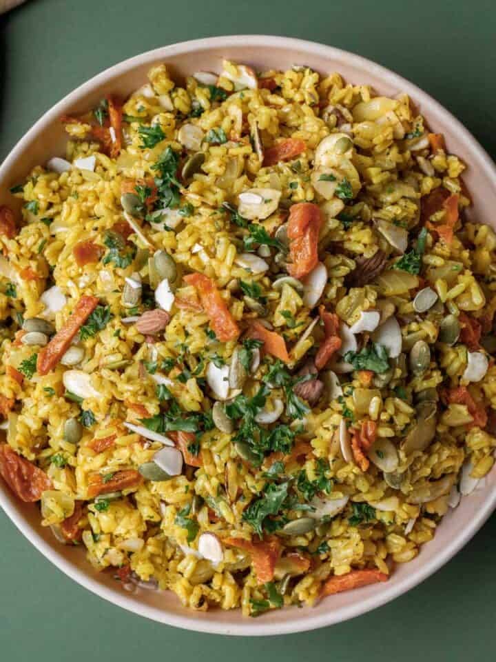 Colorful brown rice pilaf with nuts, seeds, and apricots in a bowl on a green surface.