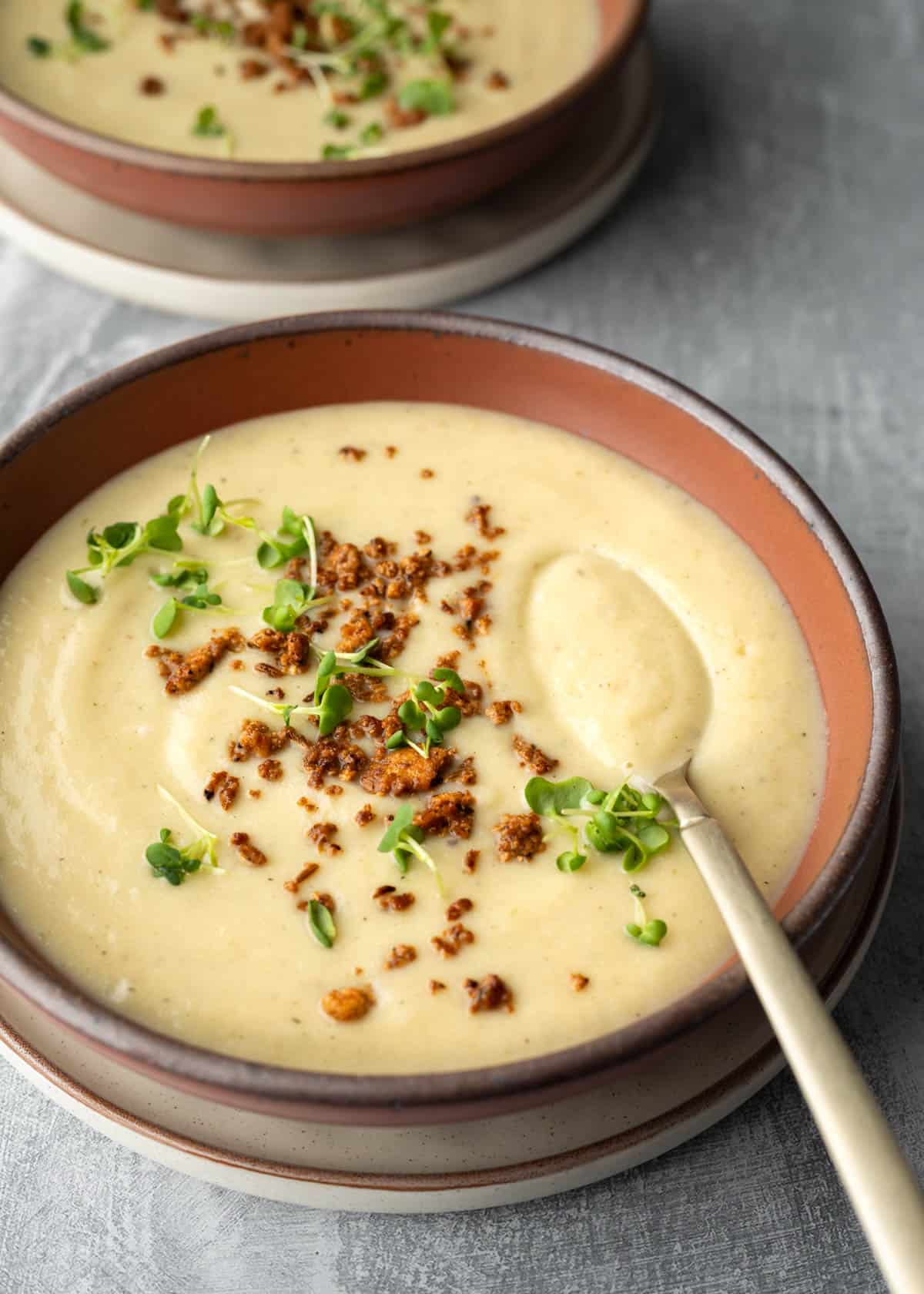 Two bowls of vegan cauliflower soup with a spoon dipping in to show the creamy consistency.